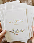 Person holding a stack of greeting cards with 'welcome little one' text and bird illustration.