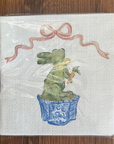 A square white napkin with a printed Easter bunny design sitting in a blue basket and holding a carrot, under a pink ribbon bow, shown wrapped in clear plastic on a wooden surface.