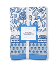 A folded blue and white floral block print tablecloth with a label indicating 'Two's Company' and 'Botanica'. It has a napkin ring attached to the folded edge.