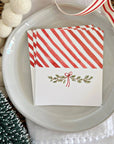 A stack of white place cards with red diagonal stripes on the top half and a small red-and-green holiday sprig motif on the bottom half, arranged on a white plate with decorative pom-poms, a ribbon, and a textured surface around it.