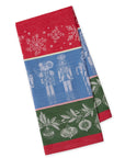 A folded holiday-themed woven towel featuring a red top section with white snowflakes, a blue middle section with white nutcracker figures, and a green bottom section with white ornaments and leaves.