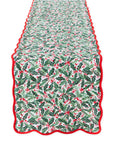 A quilted table runner with a festive holly and berry pattern in green, white, and red, finished with a scalloped red border, shown flat against a white background.