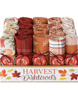 Dishtowels in autumn harvest colors, featuring various plaid, striped, and patterned cotton cloths tied with red ribbons, with pumpkin-shaped soap bars in front. The wooden box has 'HARVEST Dishtowels' printed on the front with fall leaves.