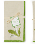 Two folded cloth napkins on a white background. Each napkin is made of natural linen with a green border, decorated with embroidered botanical motifs, and tied with a ribbon and paper tag that reads CLOTH NAPKINS.