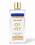 A clear rectangular glass bottle of Chez Gagne Oy Vey safety matches with a gold cap, white label, blue match heads, and visible wooden matchsticks inside, on a white background.
