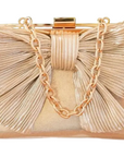 A metallic champagne gold clutch bag with a large pleated bow design, a gold chain strap, and a gold metal clasp, set against a background of green foliage and white geometric blocks.