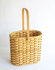 A handwoven tall gathering basket with a plywood bottom and a solid brass nameplate, featuring a natural seagrass color and a brown leather handle.