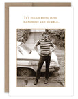 Greeting card with a black and white photo of a man standing next to a car, with text 'It's tough being both handsome and humble.'.