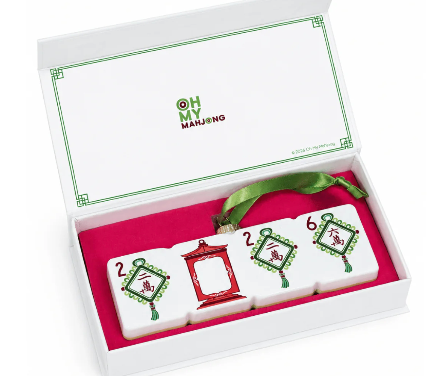 An ornament of four white Chinese mahjong tiles displayed inside an open white gift box with a pink interior and green ribbon. The lid shows the brand name 'Oh My Mahjong'.