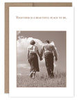 Greeting card with a black and white photo of two people holding hands in a field, with text 'Together is a beautiful place to be.'.