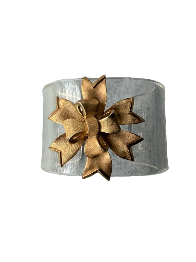 A clear cylindrical cuff bracelet featuring a large, textured metallic gold bow attached to the front, set against a white background.