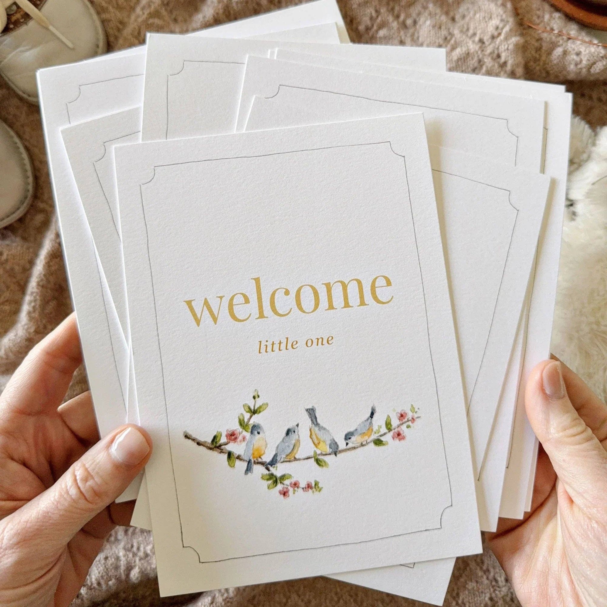 Person holding a stack of greeting cards with 'welcome little one' text and bird illustration.