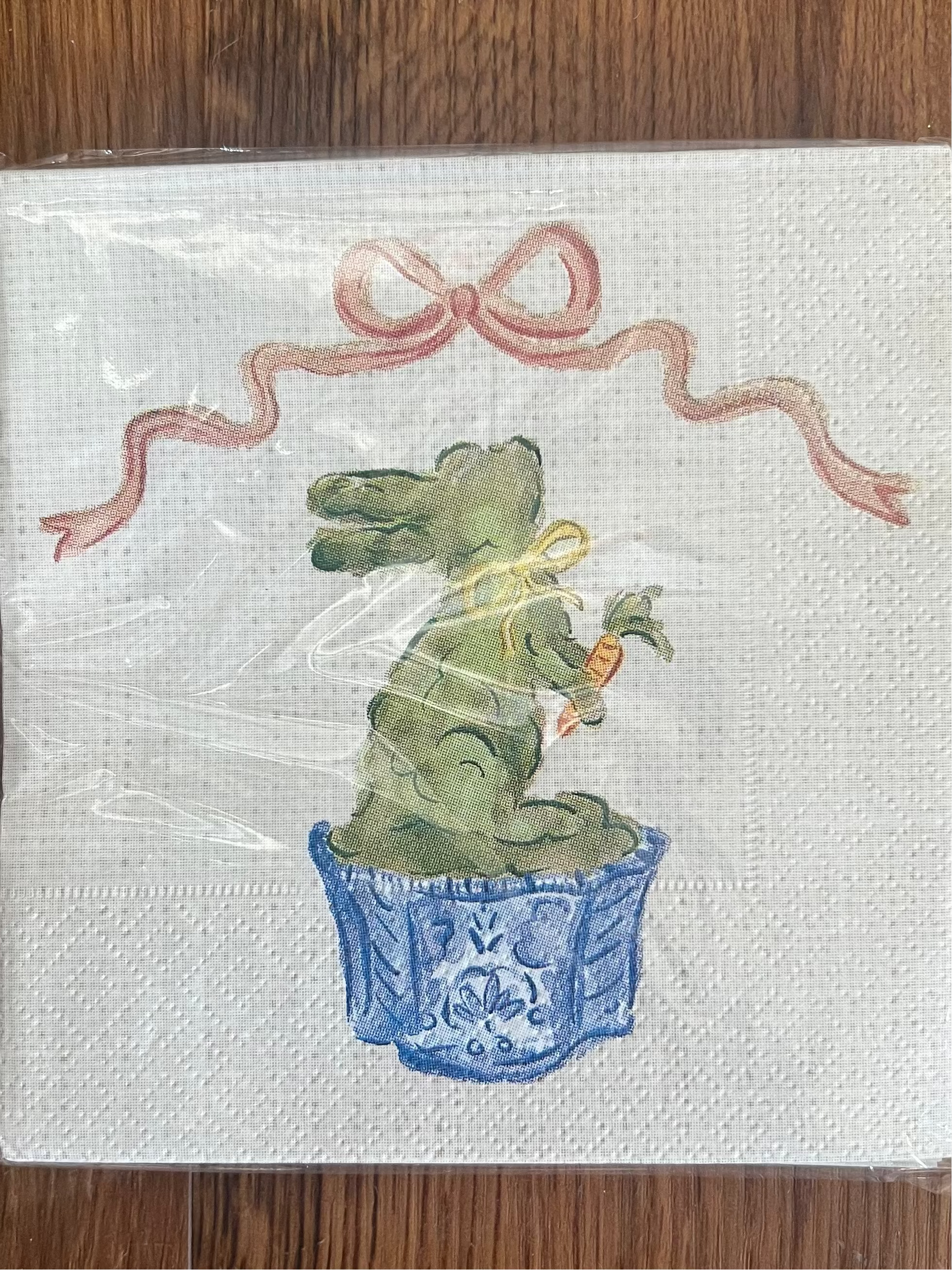 A square white napkin with a printed Easter bunny design sitting in a blue basket and holding a carrot, under a pink ribbon bow, shown wrapped in clear plastic on a wooden surface.