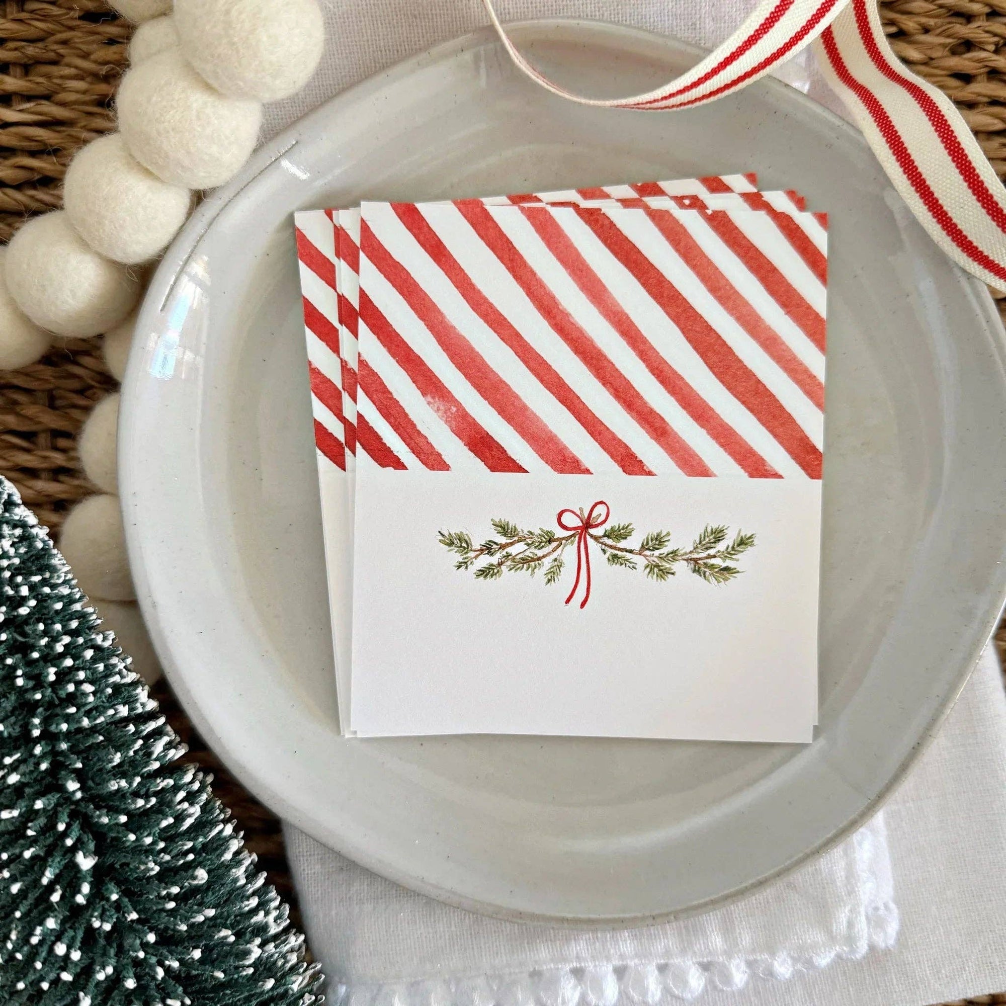 A stack of white place cards with red diagonal stripes on the top half and a small red-and-green holiday sprig motif on the bottom half, arranged on a white plate with decorative pom-poms, a ribbon, and a textured surface around it.