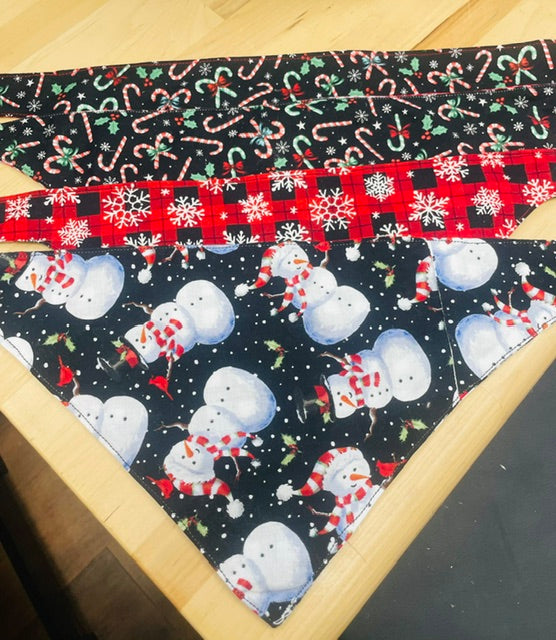 A triangular dog bandana with a Christmas theme, featuring a black background with white snowmen, red and white candy canes, and green holly accents, laid flat on a light wood surface.