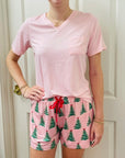 A woman wearing a light pink short-sleeve V-neck t-shirt and matching pink pajama shorts with a green Christmas tree pattern and a red drawstring, standing in front of a white paneled door.