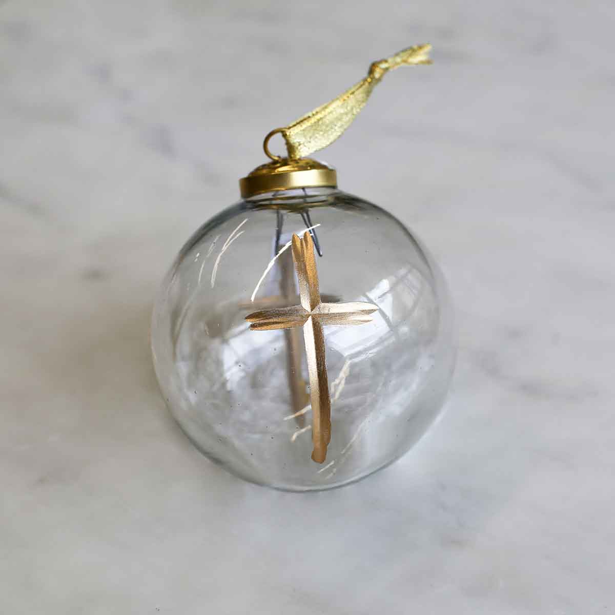 A clear glass Christmas ornament with a gold metal cap, gold hanging ribbon, and a wooden cross inside, set on a light marble surface.