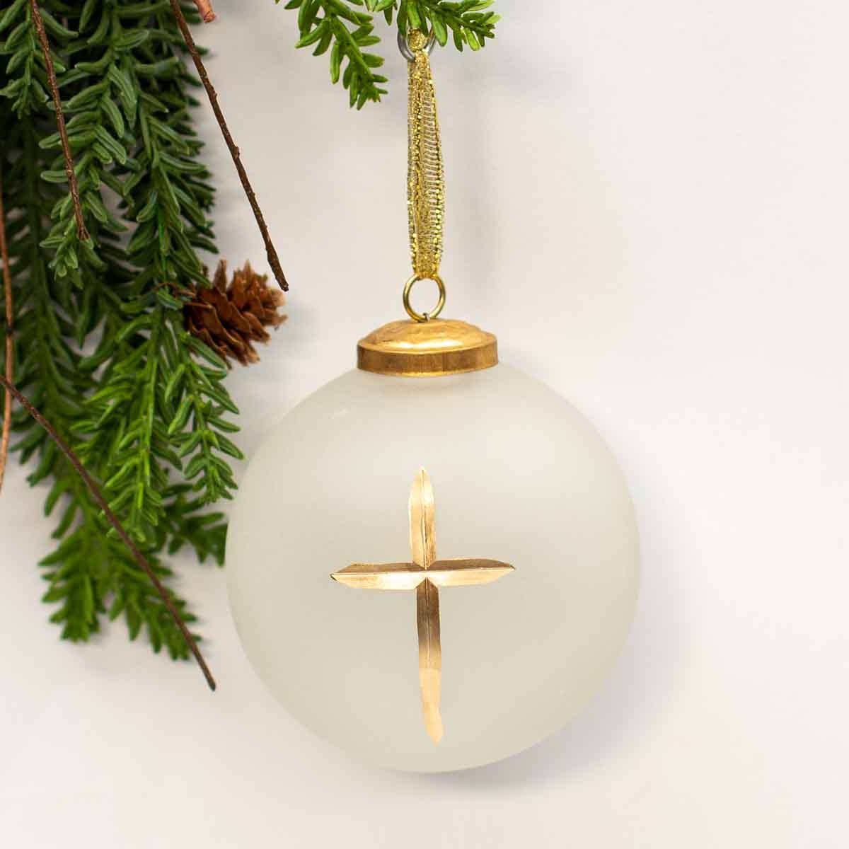 A frosted glass Christmas ornament shaped like a sphere, featuring a gold cross in the center and a gold hanging loop, styled with green pine branches against a white background.