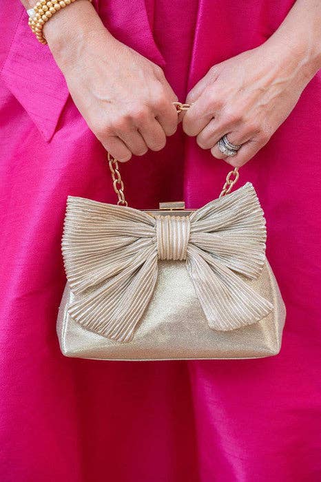 Gold clutch with a large bow detail held by a person wearing a pink dress.