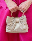 Gold clutch with a large bow detail held by a person wearing a pink dress.