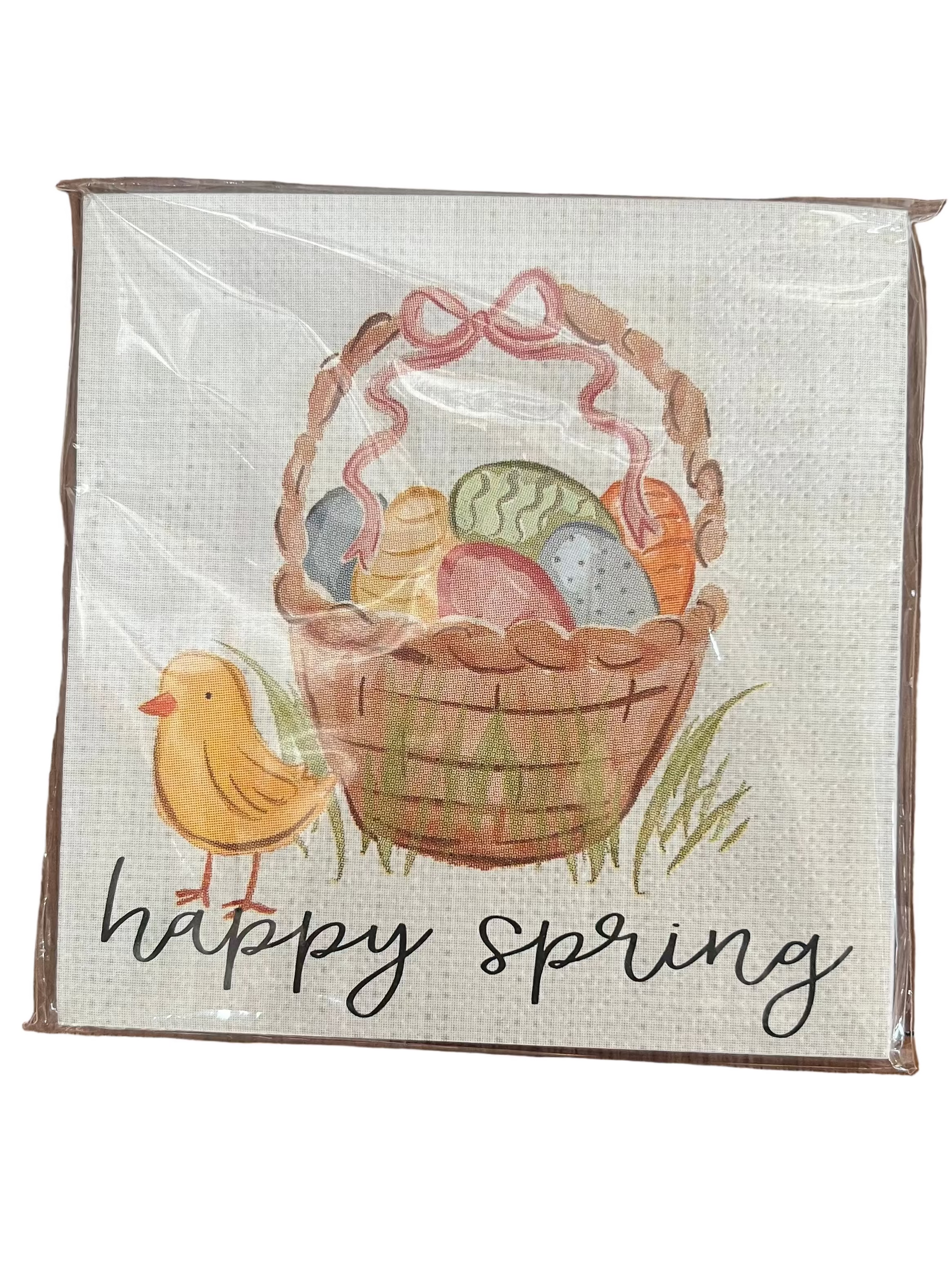 A square decorative sign or print wrapped in clear plastic, featuring an Easter basket filled with colorful eggs, a small yellow bird, and the words 'happy spring' on a light textured background.