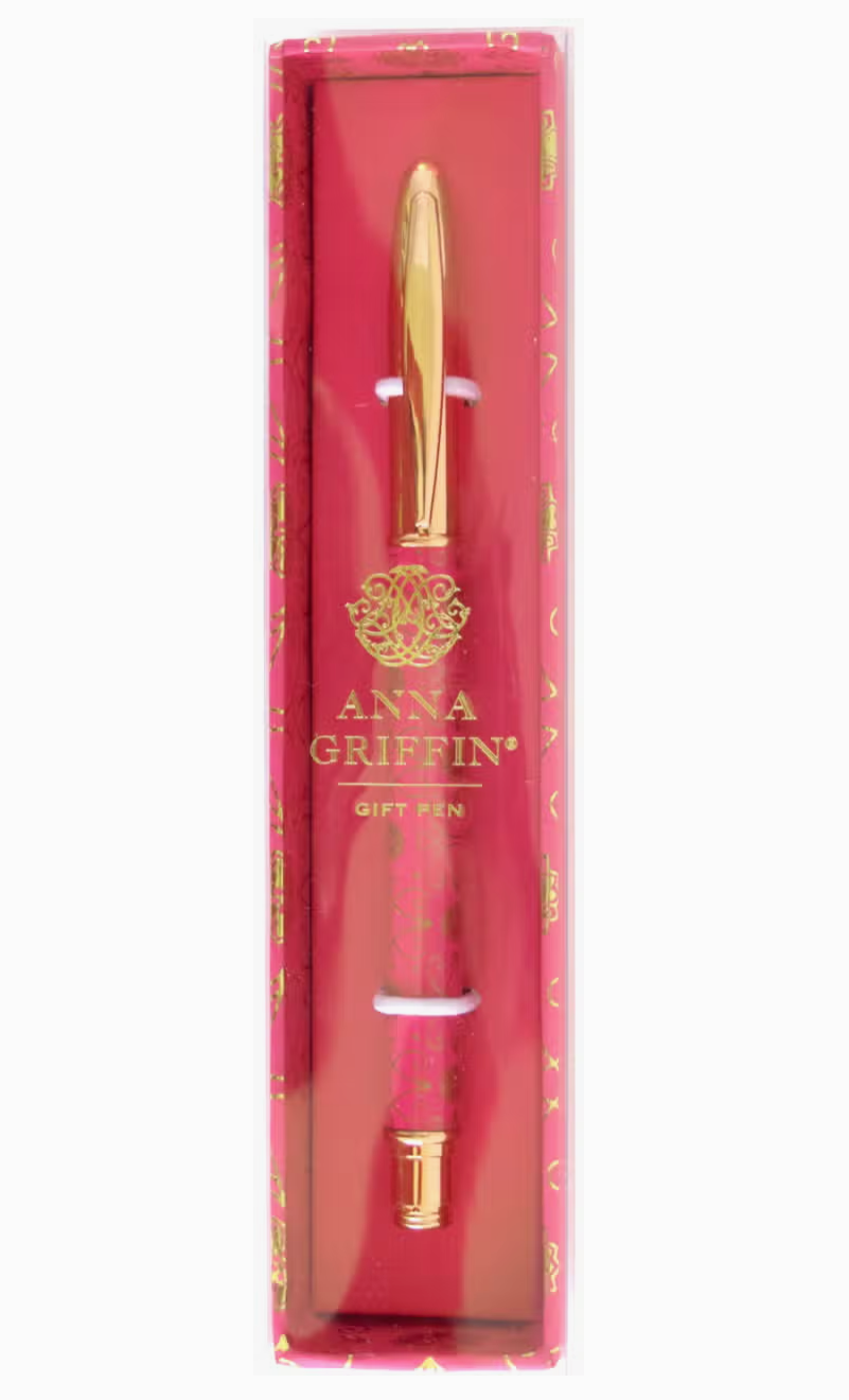 A hot pink pen with a gold cap, displayed in an upright position within its packaging. The packaging has a golden floral pattern and the brand 'Anna Griffin' is visible.