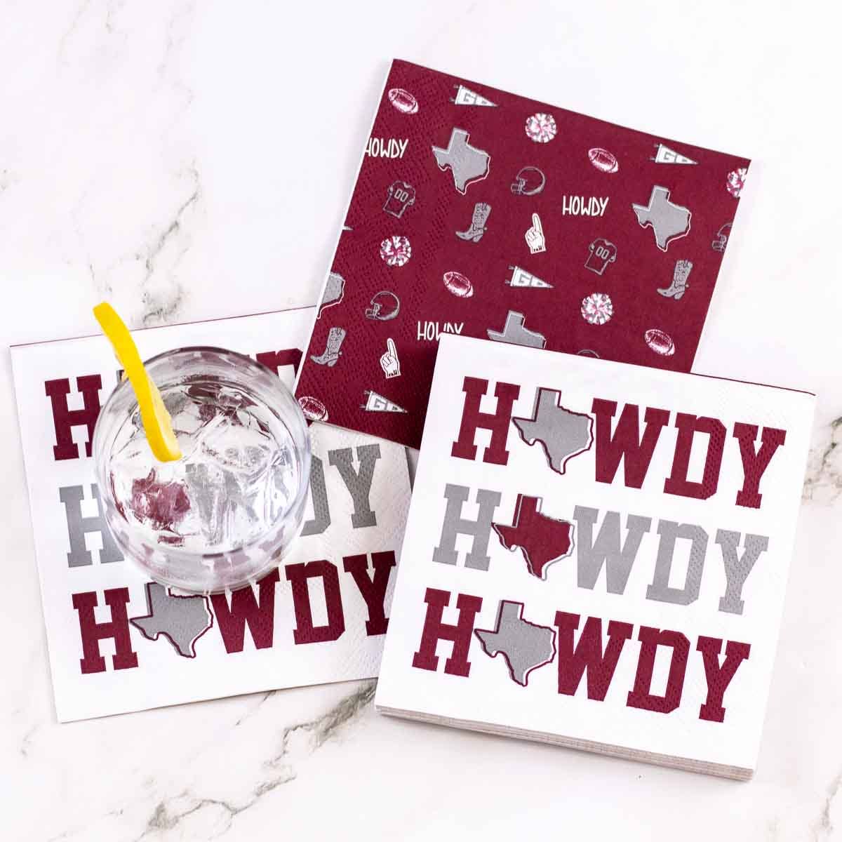 Cocktail napkins with a holiday Texas theme are arranged on a white marble surface. Napkins are white with burgundy and gray text reading 'HOWDY' and a gray silhouette of Texas, while one napkin is burgundy with small holiday icons.