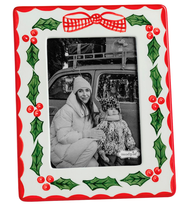 A rectangular Christmas-themed photo frame with a red border, green holly leaves, red berries, and a red bow motif on a white background. The center holds a black-and-white photo of a woman and child in winter clothing.