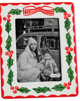 A rectangular Christmas-themed photo frame with a red border, green holly leaves, red berries, and a red bow motif on a white background. The center holds a black-and-white photo of a woman and child in winter clothing.