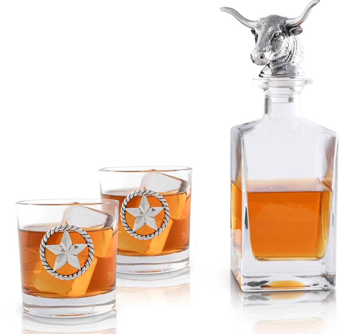 A clear glass decanter with a metal longhorn accent and a 3-D decanter stopper, accompanied by a pair of glasses, all displayed against a white background.