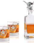 A clear glass decanter with a metal longhorn accent and a 3-D decanter stopper, accompanied by a pair of glasses, all displayed against a white background.