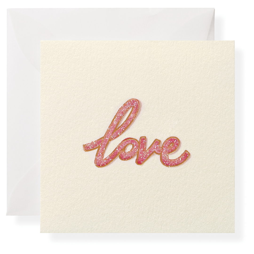 A square cream-colored greeting card with the word 'love' written in glittery pink cursive script, shown with a matching cream envelope behind it on a white background.