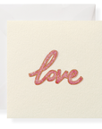 A square cream-colored greeting card with the word 'love' written in glittery pink cursive script, shown with a matching cream envelope behind it on a white background.