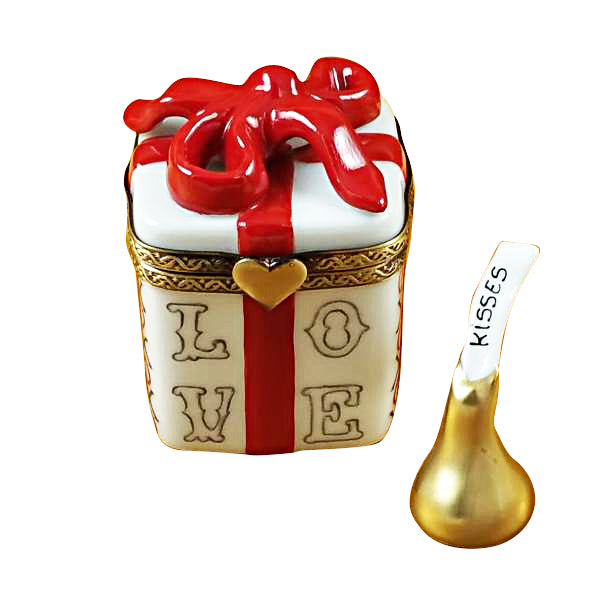 A hand-painted porcelain Limoges box in the shape of a gift box with the word 'LOVE' on the side, adorned with a red ribbon and a heart, accompanied by a golden removable element resembling a kiss.