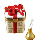 A hand-painted porcelain Limoges box in the shape of a gift box with the word 'LOVE' on the side, adorned with a red ribbon and a heart, accompanied by a golden removable element resembling a kiss.
