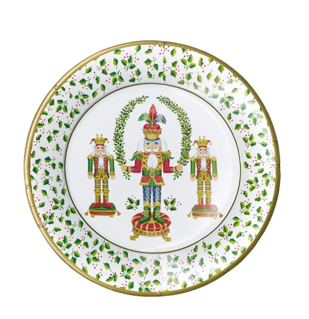 A round white paper plate featuring a festive holiday design with three nutcracker figures, green holly leaves, red berries, and a gold rim.