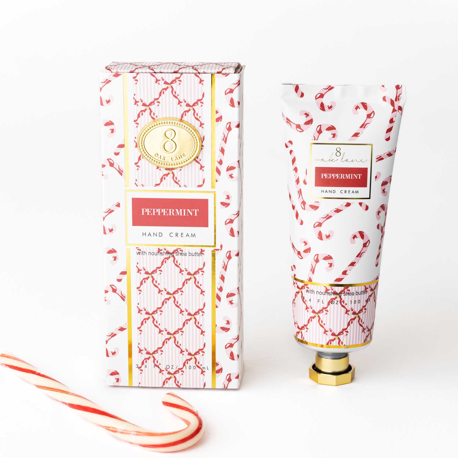 A tube and matching box of Peppermint Hand Cream, featuring a red and white candy cane and lattice pattern with gold accents, displayed on a clean white background with a candy cane prop in the corner.