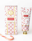 A tube and matching box of Peppermint Hand Cream, featuring a red and white candy cane and lattice pattern with gold accents, displayed on a clean white background with a candy cane prop in the corner.