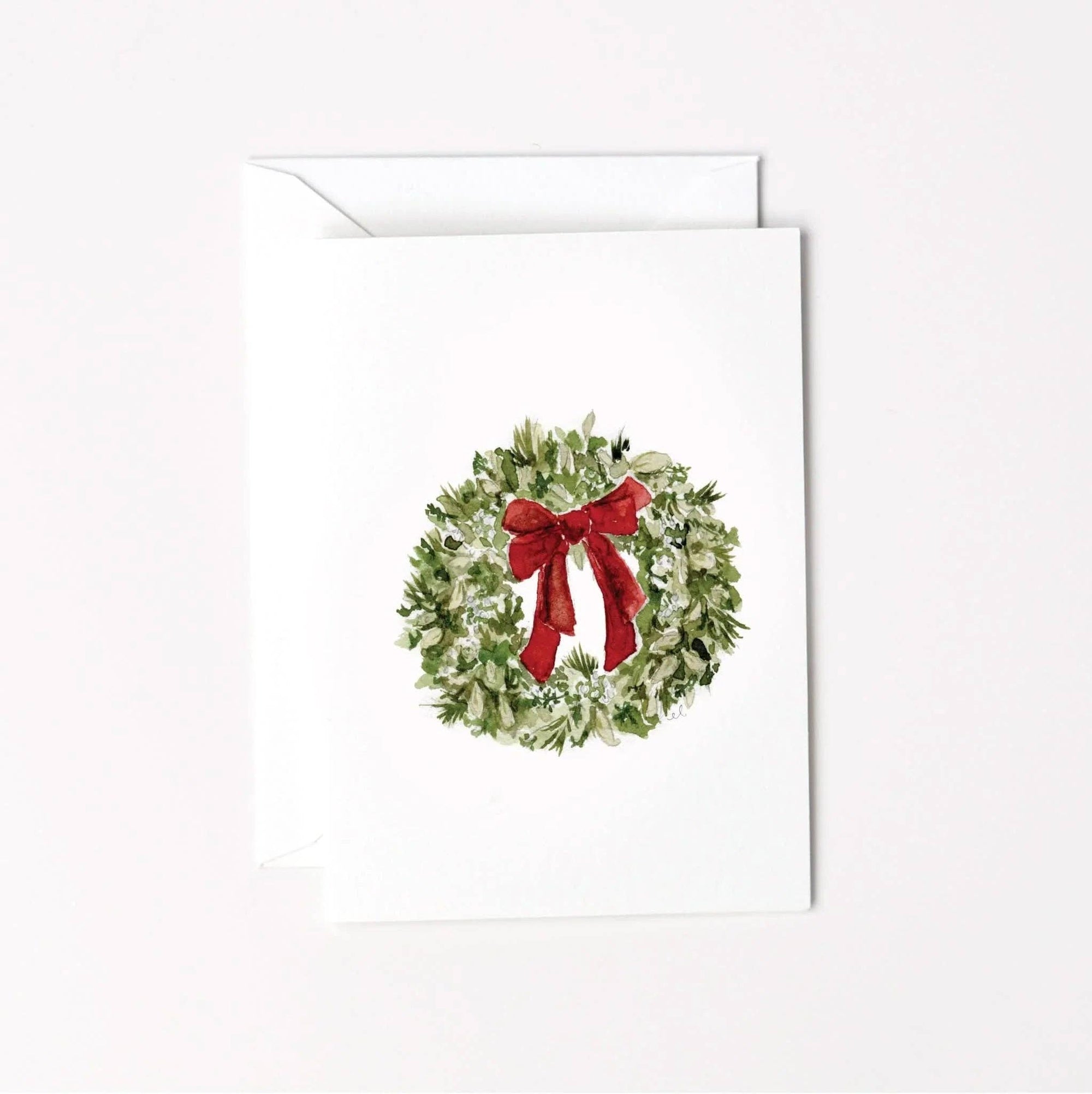 A white greeting card with a watercolor-style green wreath and red bow printed on the front, shown with a matching white envelope behind it on a light background.