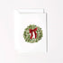 A white greeting card with a watercolor-style green wreath and red bow printed on the front, shown with a matching white envelope behind it on a light background.