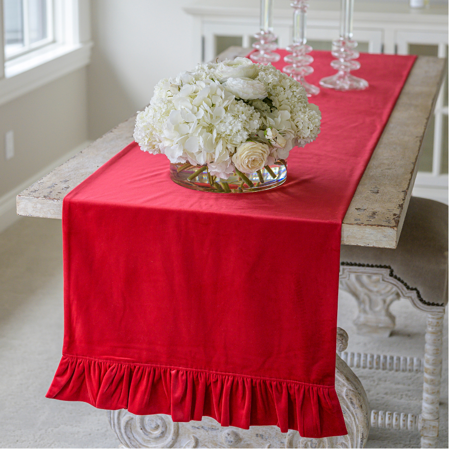 A red velvet table runner with a ruffle detail, laid out on a table with a floral arrangement in the center.