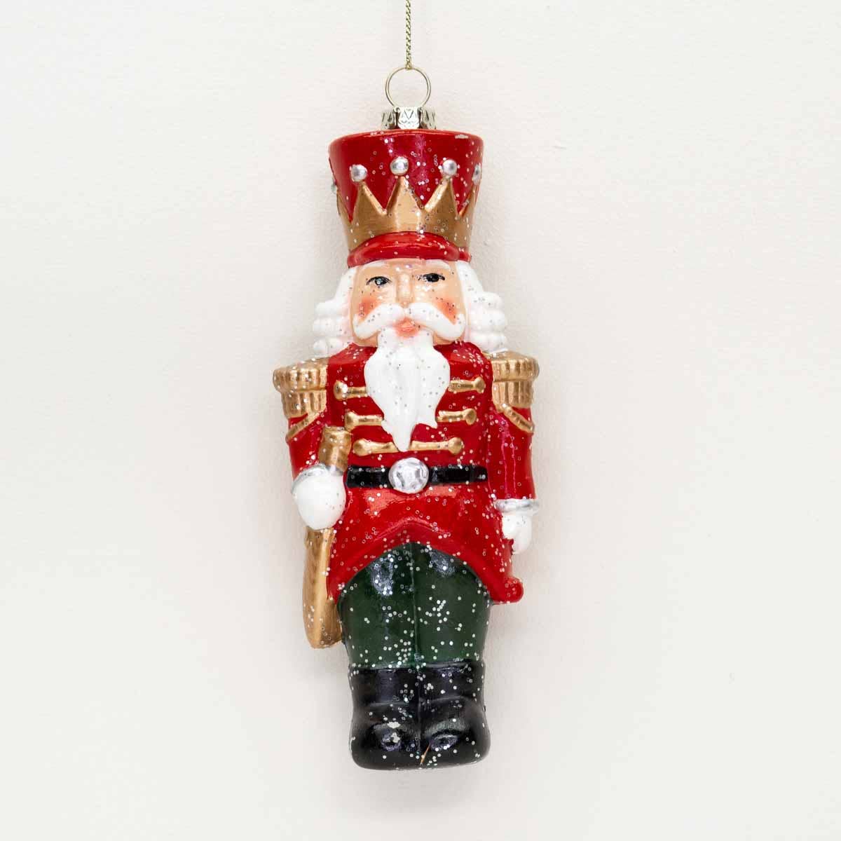 A classic Christmas nutcracker ornament hanging against a plain white background. The ornament features a red coat with gold trim, a red hat with gold accents, a white beard, and green and black pants.