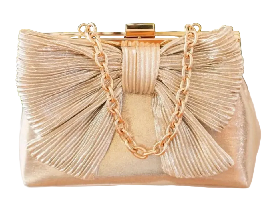 A metallic champagne gold clutch bag with a large pleated bow design, a gold chain strap, and a gold metal clasp, set against a background of green foliage and white geometric blocks.