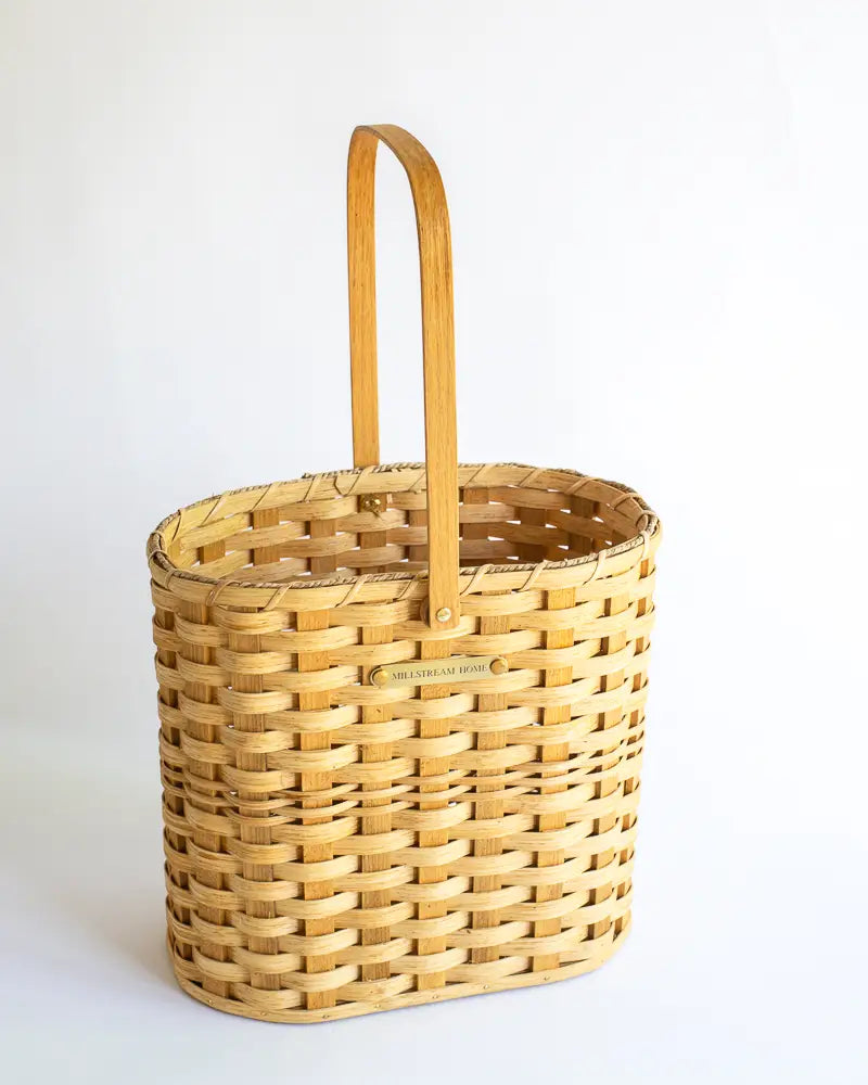 A handwoven tall gathering basket with a plywood bottom and a solid brass nameplate, featuring a natural seagrass color and a brown leather handle.