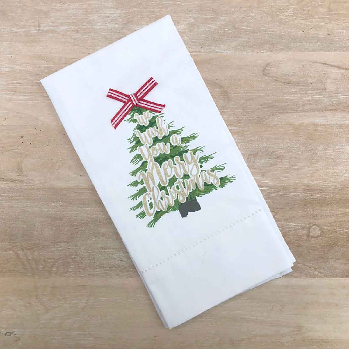 A white kitchen towel with a 'Merry Merry Christmas' tree design, displayed diagonally on a light wood grain surface.