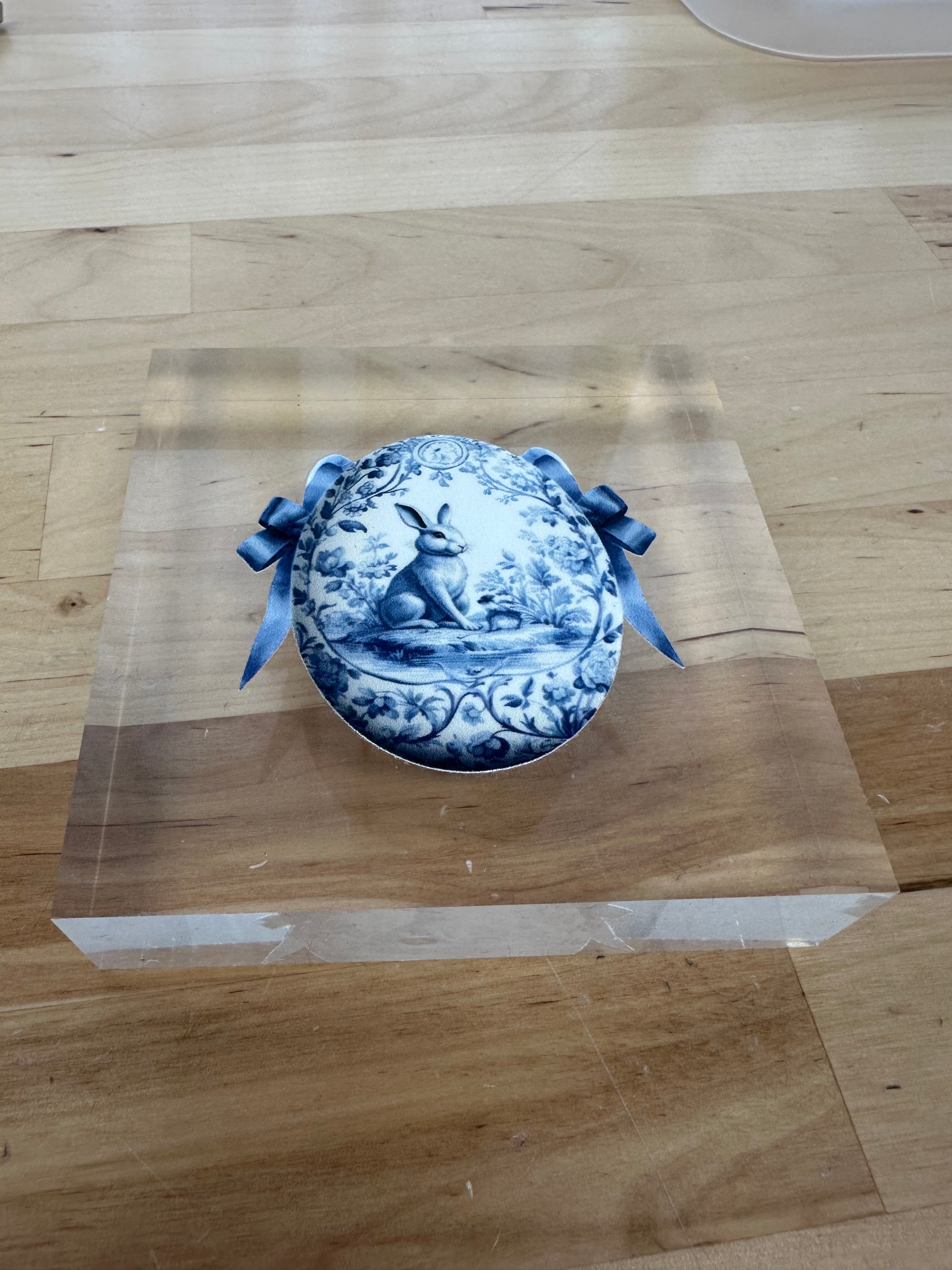 A blue and white chinoiserie decorative egg placed inside a clear acrylic block, with blue ribbons on the corners of the block.